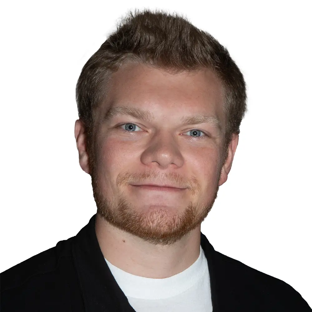 A young man with a beard smiles while wearing a black polo shirt and a white undershirt, against a plain white background.